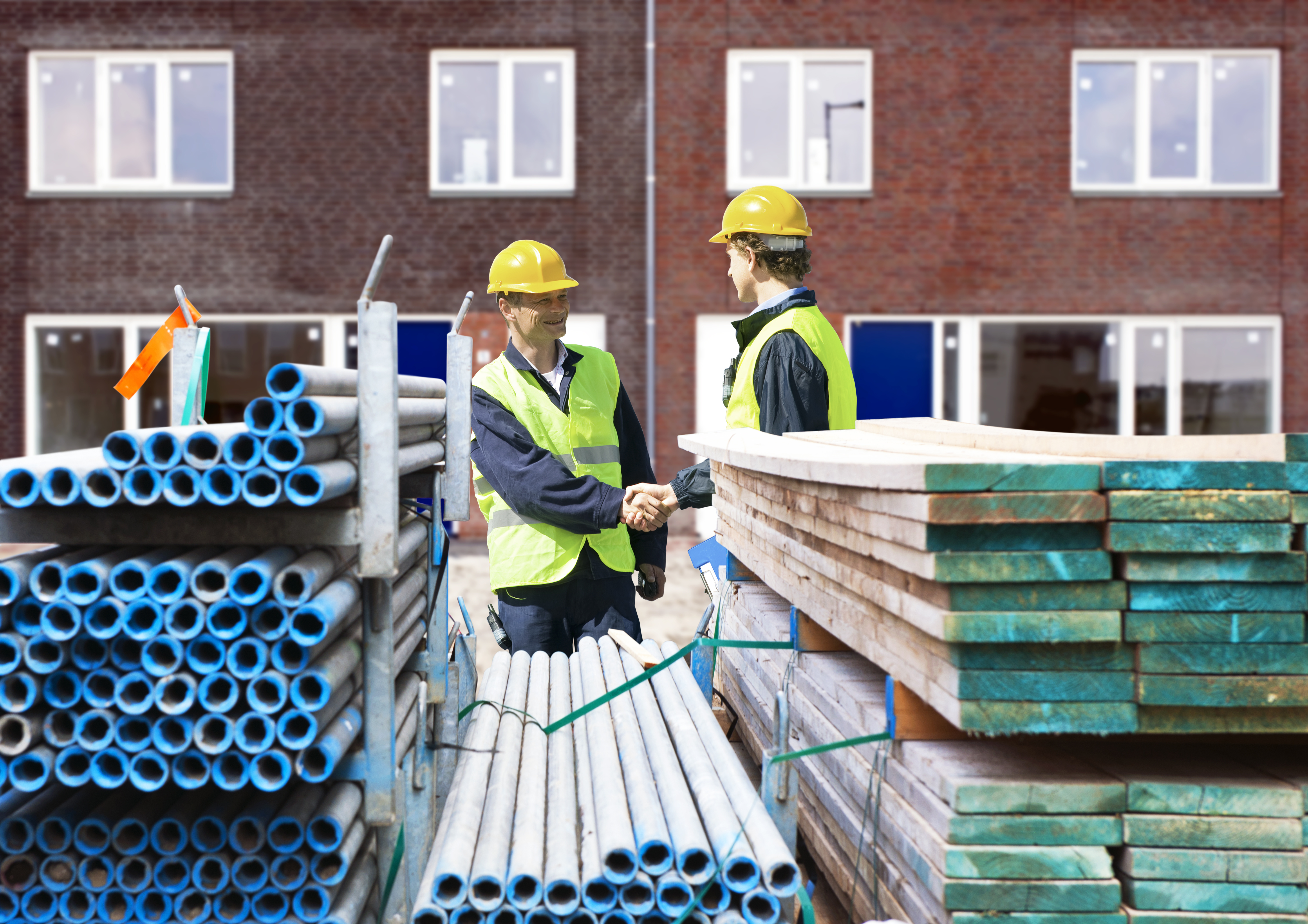 Two building contractors shaking hands behind stacks of scaffolding material, in front of a newly completed residential building complex