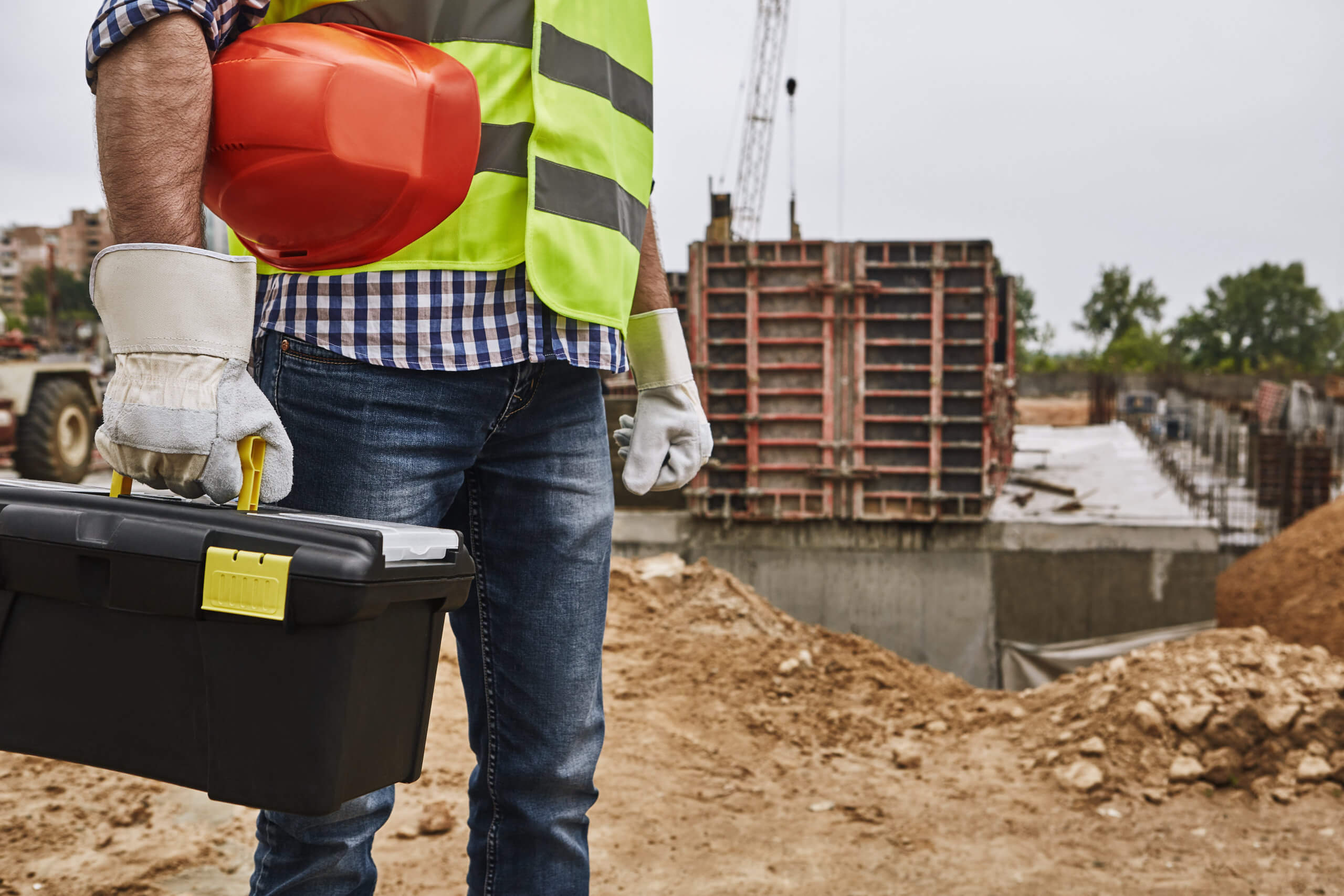 contractor Ready to work! Cropped image of a builder in working uniform holding red helmet and carrying toolbox while standing at construction site. Building concept. Protective gear. Construction concept